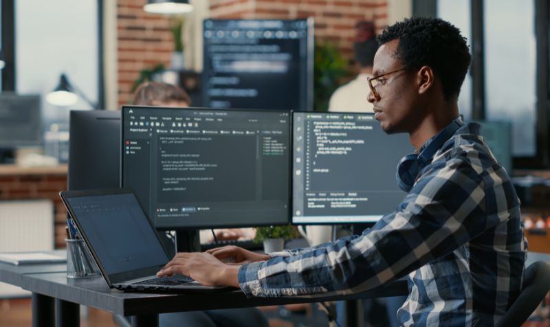 Developer using laptop to write code sitting at desk with multiple screens parsing algorithm in software agency. Coder working on user interface using portable computer. 